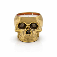 A gold skull-shaped candle holder with a glossy metallic finish, featuring a visible orange wax interior, set against a clean white background.