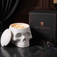 A white skull-shaped candle with three lit wicks sits beside its black branded box on a dark marble surface. The lid rests against the left side of the skull, and a black rose and small stones are placed nearby.