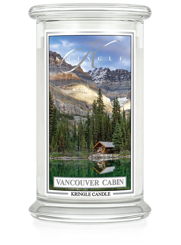 Unlit 2-wick candle in a clear jar. Label shows a cabin by a lake, surrounded by trees and mountains under a blue sky.