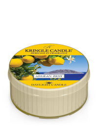 Kringle Candle Aegean Zest Daylight Candle – A small, cream-colored candle in a plastic cup with a vibrant lid design featuring fresh lemons, white blossoms, and a scenic ocean view in the background. The label includes ‘Kringle Candle - Heritage in Fragrance,’ the fragrance name ‘Aegean Zest,’ and ‘Daylight Candle’ branding in yellow and white text.