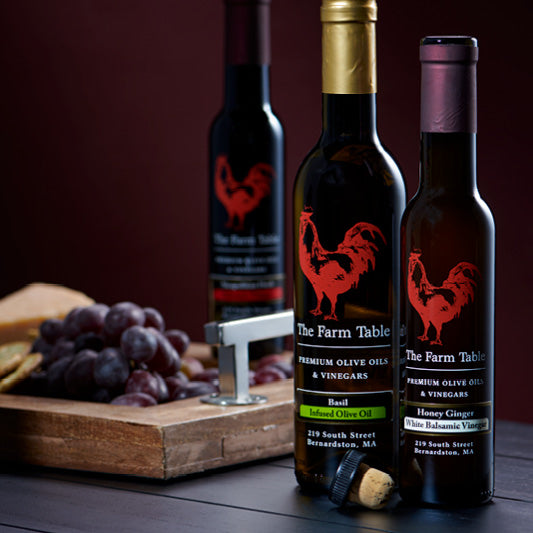 Three bottles of The Farm Table premium olive oils and vinegars displayed on a dark wood surface. The labels feature a red rooster logo and flavors such as Basil Infused Olive Oil and Honey Ginger White Balsamic Vinegar. A wooden charcuterie board with grapes, crackers, and a bottle opener sits beside the bottles, with a rich, warm backdrop completing the gourmet setting.