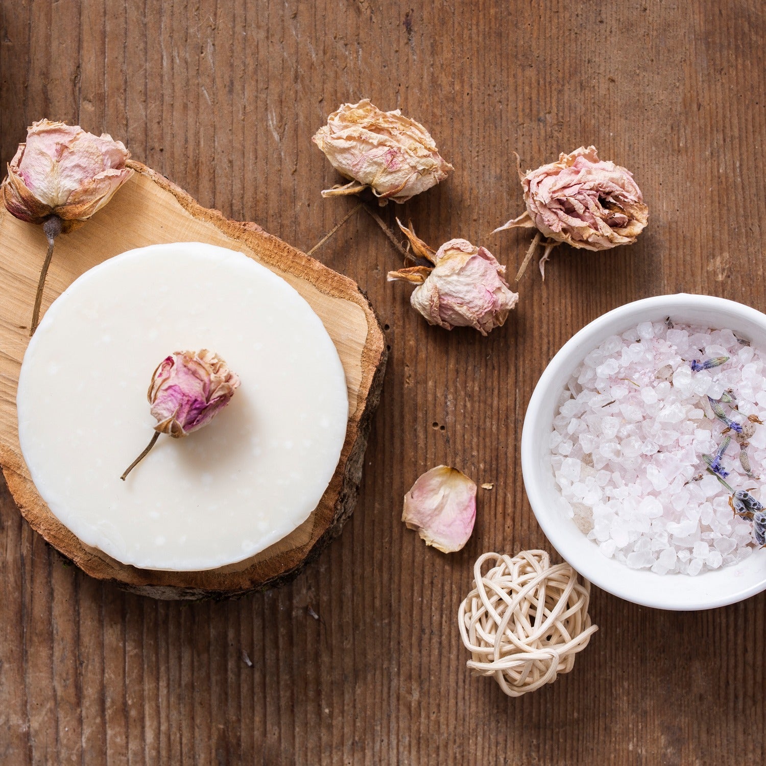 Candle with dried flowers on a wooden surface, next to a bowl of bath salts.