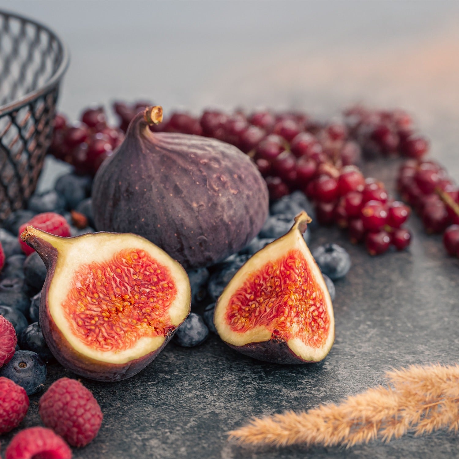 Figs with a halved fig showing red flesh, surrounded by grapes and berries on a dark surface.