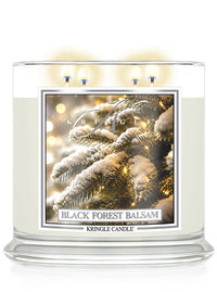 A large Kringle Candle jar labeled 'Black Forest Balsam' with three lit wicks at the top, shown on a clean white background. The front label features a festive image of snow-covered pine branches with warm string lights.