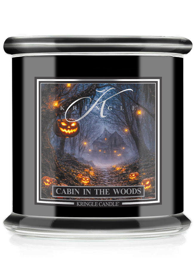 A Kringle Candle jar candle with a black body and lid, featuring a Halloween-themed label that reads 'Cabin in the Woods' with a haunted forest scene and glowing jack-o'-lanterns.