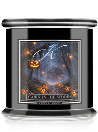 A Kringle Candle jar candle with a black body and lid, featuring a Halloween-themed label that reads 'Cabin in the Woods' with a haunted forest scene and glowing jack-o'-lanterns.