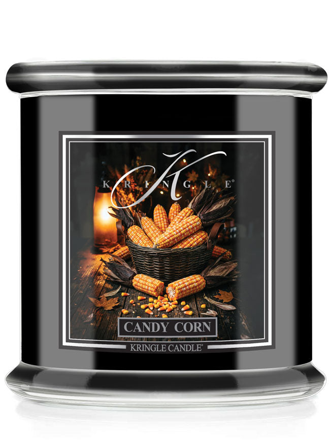 A black glass jar candle with a silver lid and a decorative front label reading 'Candy Corn' by Kringle Candle. The label shows a warm autumn still life with corn cobs, leaves, and a candle flame.