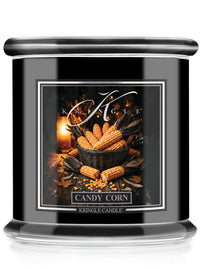A black glass jar candle with a silver lid and a decorative front label reading 'Candy Corn' by Kringle Candle. The label shows a warm autumn still life with corn cobs, leaves, and a candle flame.