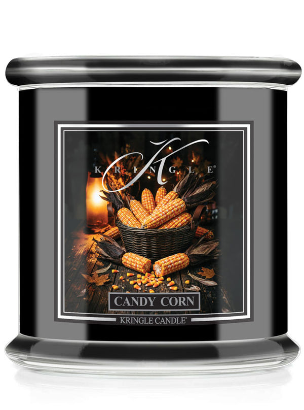 A black glass jar candle with a silver lid and a decorative front label reading 'Candy Corn' by Kringle Candle. The label shows a warm autumn still life with corn cobs, leaves, and a candle flame.