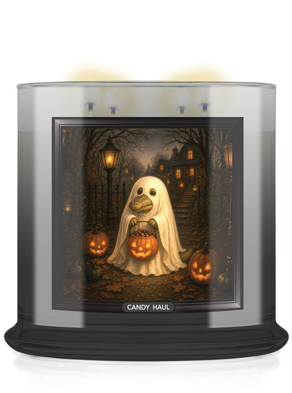 A large scented candle in a clear glass jar with a black base, featuring a Halloween-themed printed label. The label shows a ghost alligator holding a glowing jack-o'-lantern in a spooky forest scene with pumpkins and houses.
