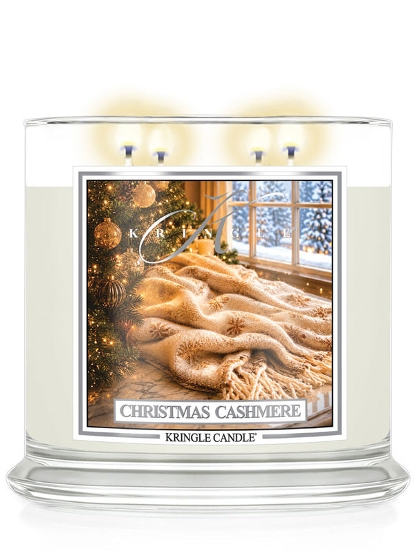A Kringle Candle 'Christmas Cashmere' scented candle in a clear glass jar with three lit wicks, featuring a warm-toned label depicting a cozy winter scene with a cashmere blanket, Christmas tree, and snowy window.
