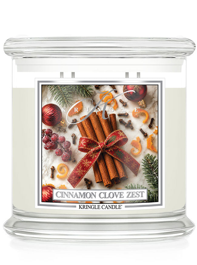 A Kringle Candle jar candle with a clear glass body and white wax, featuring a festive holiday label that reads 'Cinnamon Clove Zest.' The label shows cinnamon sticks tied with a red bow, evergreen branches, berries, cloves, and citrus slices on a light background.