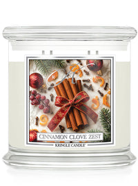 A Kringle Candle jar candle with a clear glass body and white wax, featuring a festive holiday label that reads 'Cinnamon Clove Zest.' The label shows cinnamon sticks tied with a red bow, evergreen branches, berries, cloves, and citrus slices on a light background.