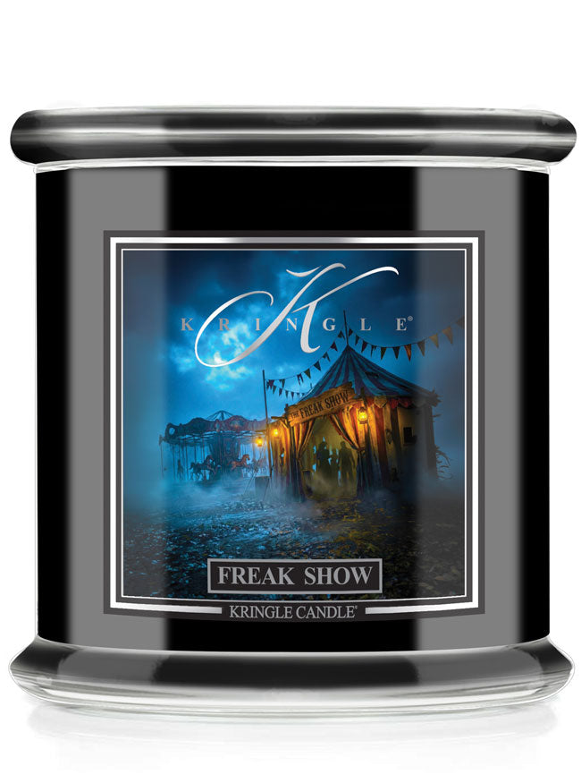 A Kringle Candle Halloween jar candle with a black body, featuring a front label that reads 'FREAK SHOW' and shows a blue-toned circus tent scene under a moonlit sky.