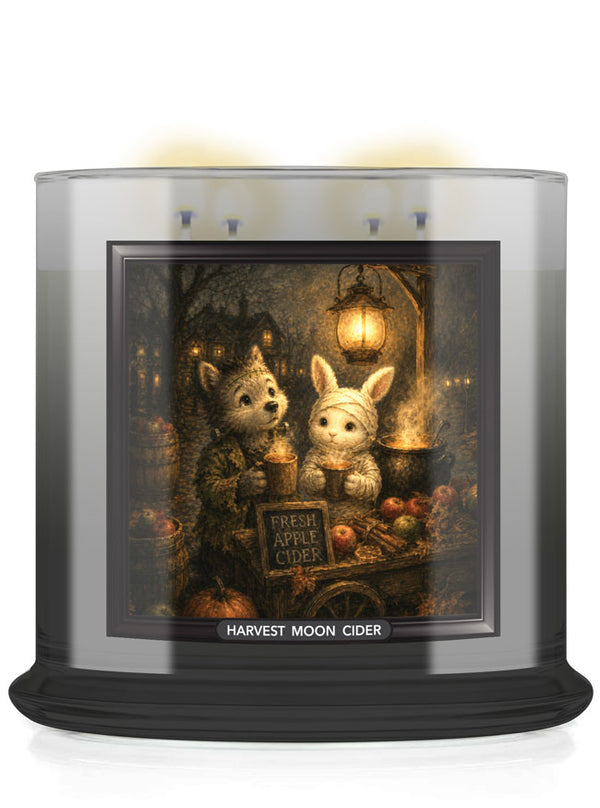 A large jar candle labeled 'Harvest Moon Cider' with a dark base and a clear top, featuring a warm illustrated label showing two cute animals, including a rabit, around a cider table at night.