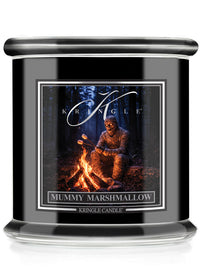 A black glass jar candle featuring a front label for Kringle Candle's 'Mummy Marshmallow' scent. The label shows a mummy sitting by a campfire in a dark forest.