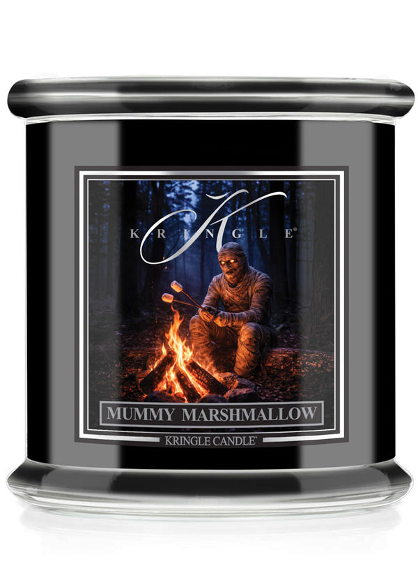 A black glass jar candle featuring a front label for Kringle Candle's 'Mummy Marshmallow' scent. The label shows a mummy sitting by a campfire in a dark forest.