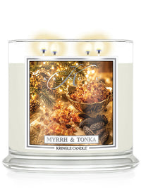 A three-wick Kringle Candle in a clear glass jar with cream-colored wax, featuring a festive 'Myrrh & Tonka' label with warm holiday imagery of spices and lights.