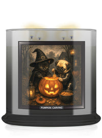 A large scented candle in a clear glass jar with a black base, featuring a Halloween-themed illustration on the front label of a black cat and a pug dog carving a jack-o'-lantern. The label reads 'PUMPKIN CARVING'.