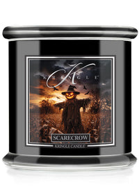 A Kringle Candle jar candle with a black body and lid, featuring a front label for the 'Scarecrow' scent with a spooky illustration of a scarecrow in a field at sunset.