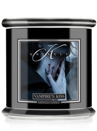 A Halloween Kringle Candle jar candle labeled 'Vampire's Kiss' with a black body, clear glass base, and a front label showing a moody portrait of a hand near a neck with red bite marks.