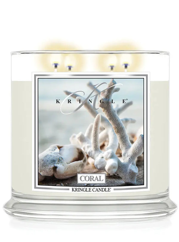Lit Kringle Candle in Coral scent with glowing flames and label showing white coral on a beach background.