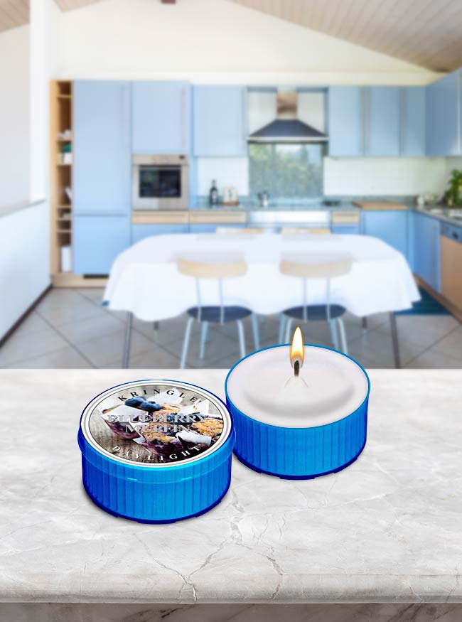 Photo of Kringle Candle's Blueberry Muffin Scented Candle DayLight that is lit and sitting on a kitchen counter, with a label that shows 2 blueberry muffins wrapped in parchment paper.
